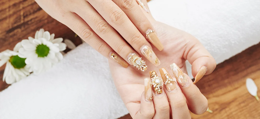Long nude coffin nails with gold foil, crystals and 3D floral art resting on a white salon towel